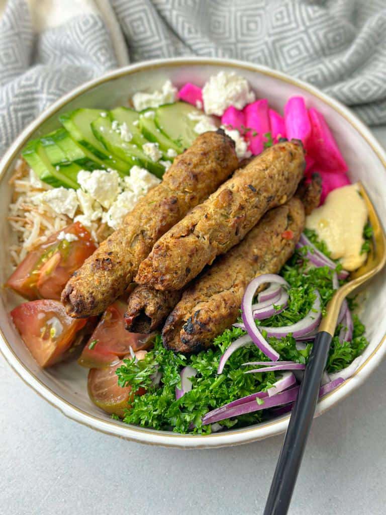A rich bowl filled with fresh parsley salad, chicken kabobs, juicy veggies, creamy hummus, and vermicelli rice.