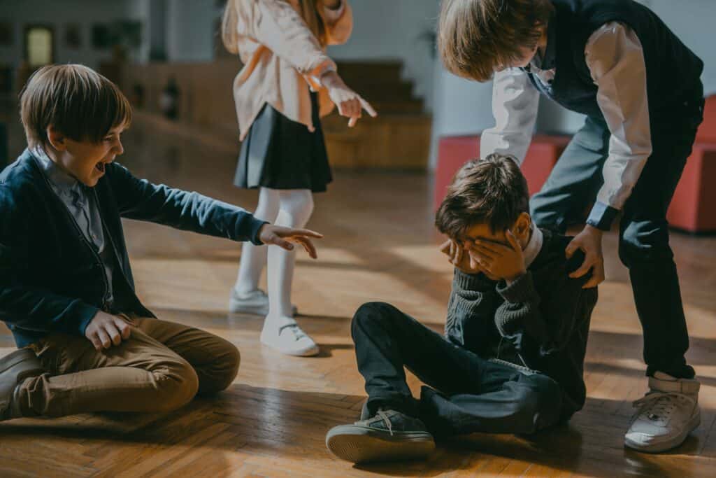 Two boys and one girl bullying a a little boy in school. The bullied boy sitting legs crisscrossed covering his face and crying.