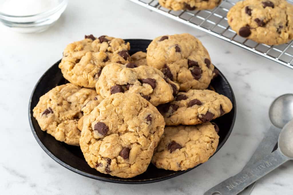 A plate of chocolate chip oatmeal cookies on a white kitchen counter with measuring spoons. Chewy chocolate chip oatmeal cookies on a white counter are visually appealing and satisfying, with a delightful texture, and a must-have for cookie lovers.