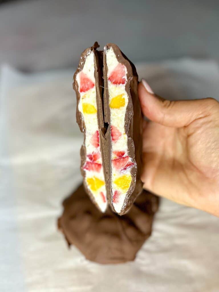 Close photo of two halves of a chocolate covered bar, showing the inside filling of yogurt, strawberries, and mango. One whole chocolate bar appears blurry in the back.
