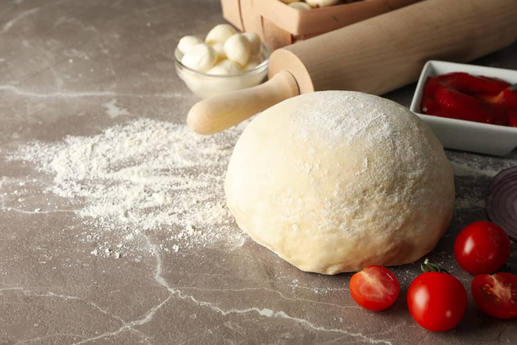 a floured surface with a pizza dough surrounded with cherry tomatoes, pizza sauce, and mozzarella cheese balls.