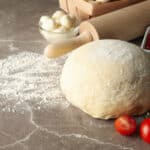 a floured surface with a pizza dough surrounded with cherry tomatoes, pizza sauce, and mozzarella cheese balls.