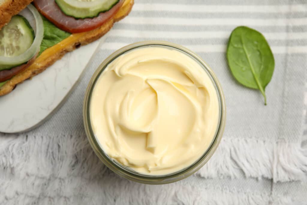 Jar of delicious mayonnaise near fresh sandwich on table, flat lay