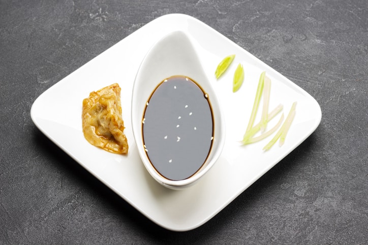 soy dressing in porcelain bowl, served with boiled gyoza dumpling and chopped leek on white plate.