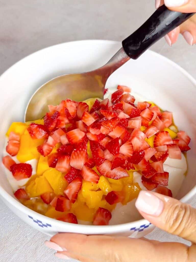 In this photo, a person is using a spoon to mix a combination of chopped mangoes and strawberries with yogurt.