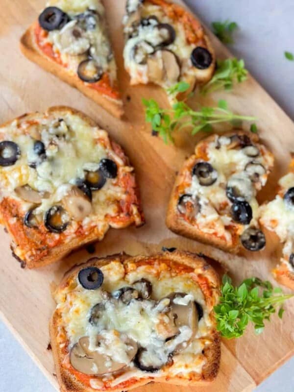 a top view of pieces of pizza toast recipe with savory toppings like cheese, mushrooms, olives, and pizza sauce are served on a wooden board