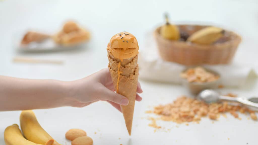 Close up view of a girl holding peanut butter banana ice-cream cone with blurred peanut, banana and topping on white table background