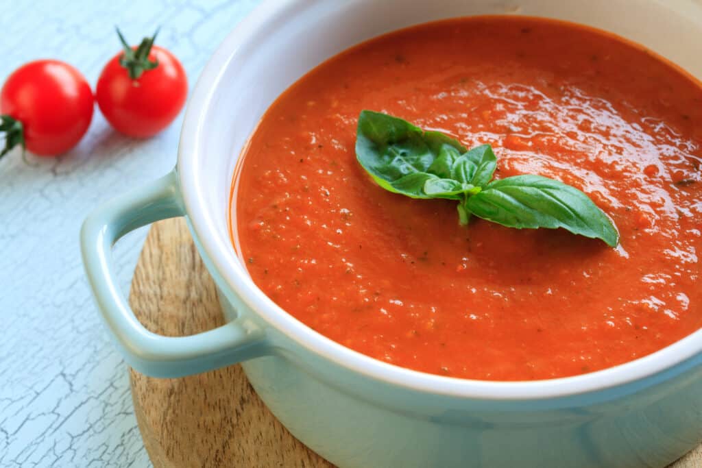 Tomato soup in a white bowl decorated with fresh basil leaves on top. Two fresh tomatoes appear in the background.