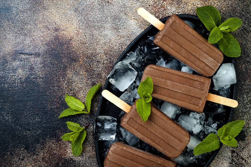 Four chocolate popsicles on a bed of ice cubes garnished with green mint leaves.