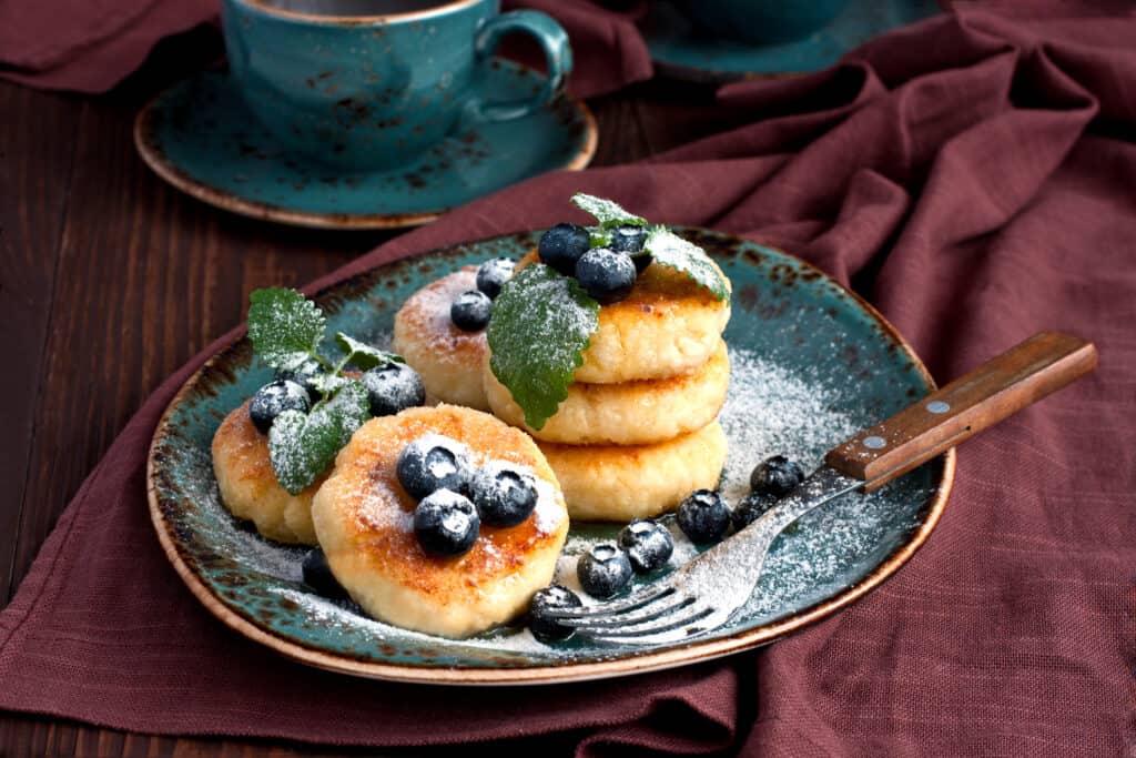 A mouth-watering photo of Cottage Cheese Pancakes topped with fresh berries and powdered sugar, served on a dark blue plate with a fork.