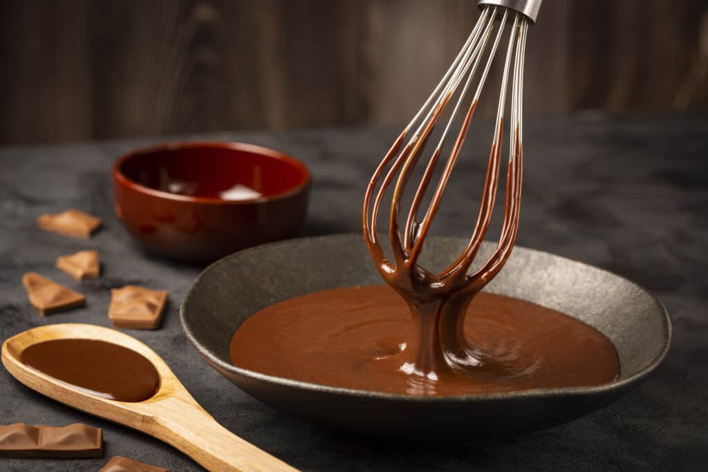 A whisk emerges from a black pot filled with luscious chocolate ganache. Accompanied by scattered chocolate cubes and a wooden kitchen spoon laden with more chocolate.