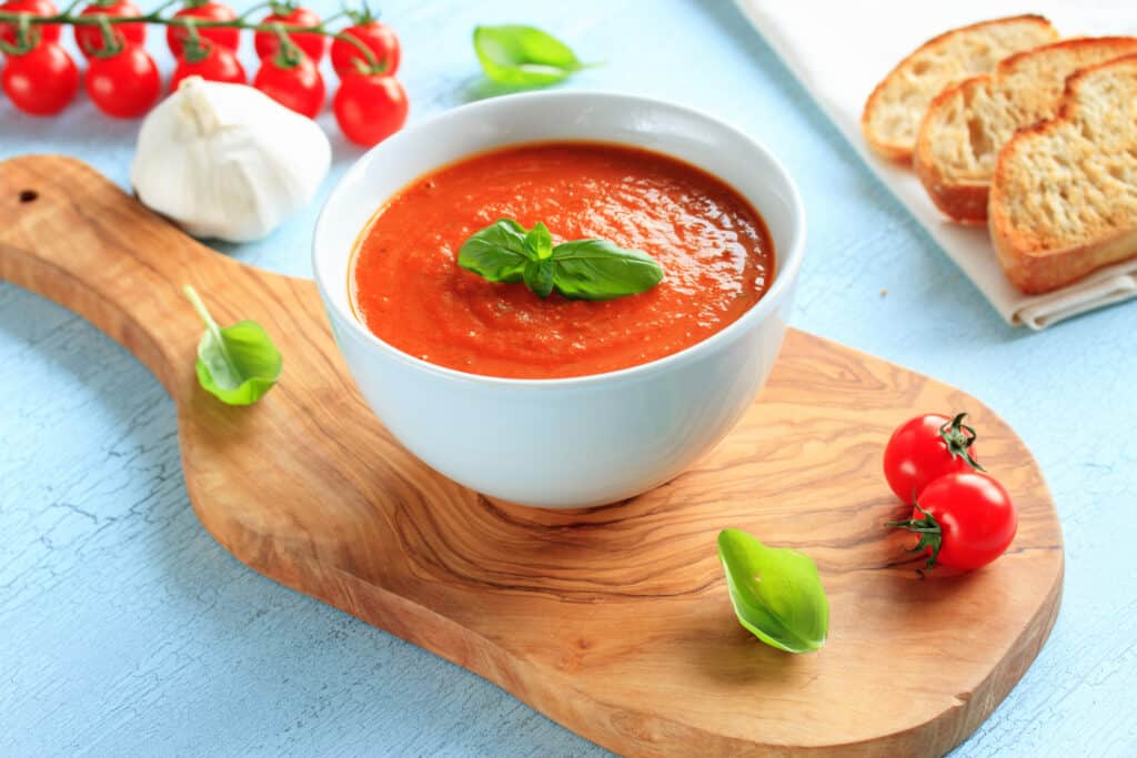 Tomato soup is set in a white, ceramic blow and decorated with basil leaves. The bowl is set on a large wooden platter. Three pieces of toasted bread appear in the back of the picture along with tomatoes and garlic.