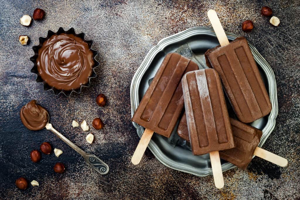 4 Nutella popsicles in a plate with a bowl and spoon of hazelnut spread on the side and chunks of hazelnuts. 