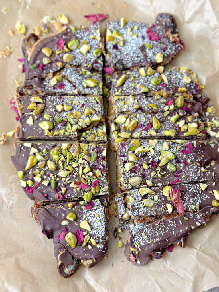 An enticing photograph captures a delectable dessert of date bark with chocolate, beautifully decorated with chopped peanuts, pistachios, coconut shreds, and dried rose petals.