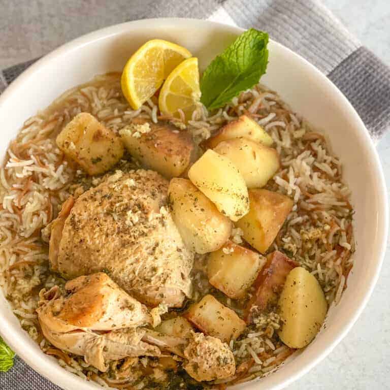 a close up of a white bowl with lemon mint garlic chicken stew and potatoes on a bed of rice decorated with lemon wedges and fresh mint