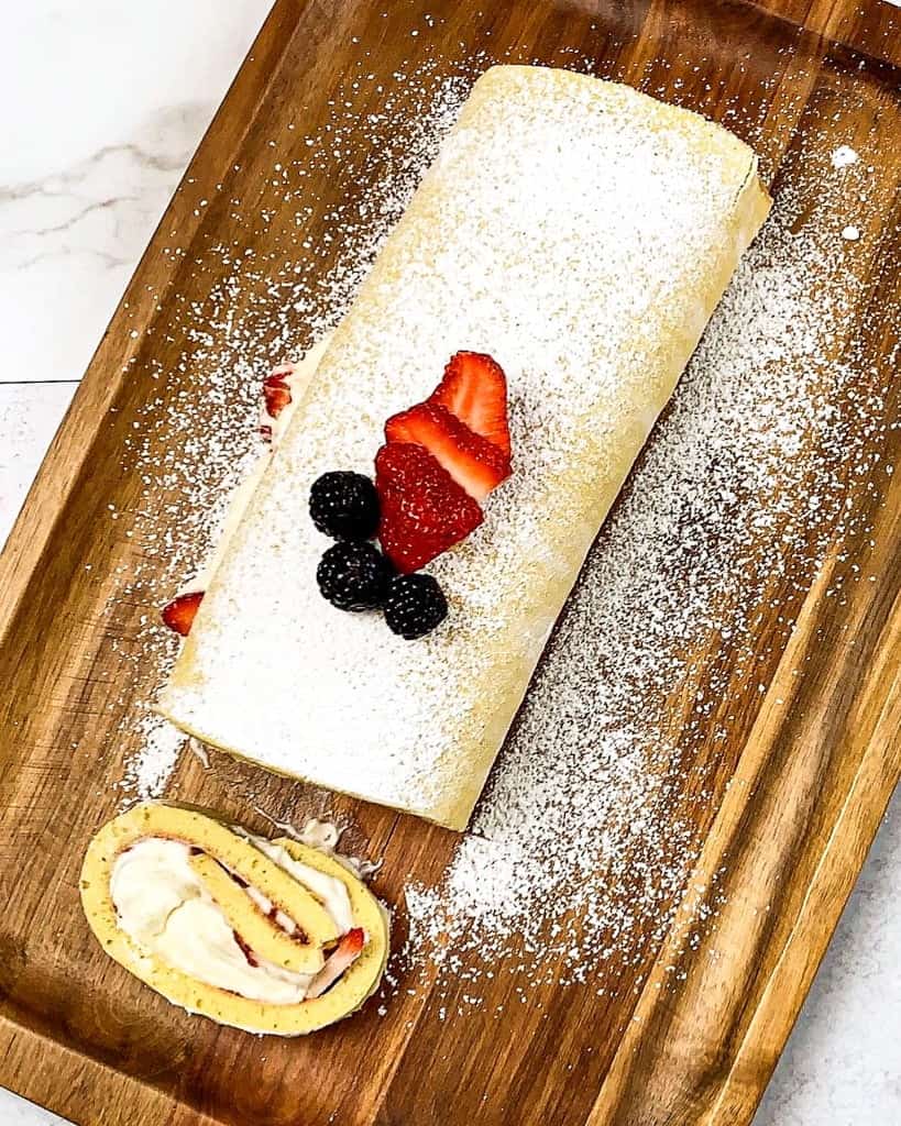 A wooden plate serving Strawberry Cheesecake Roll decorated with sprinkle of powder sugar, slices of strawberry and black berries