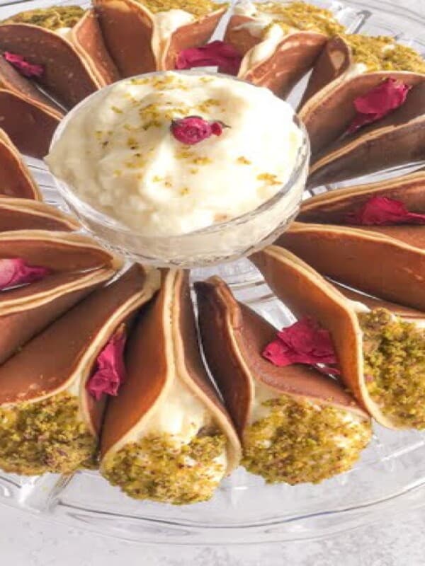 A serving plate of Middle Eastern pancakes stuffed with ashta and topped with ground pistachios with a bowl of ashta in the middle