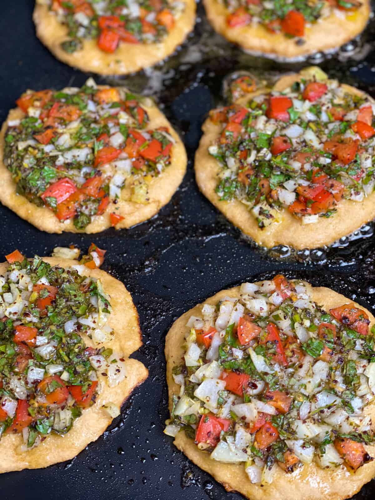 Mini veggie pizza pies topped with a mix of tomatoes, onions, sumac, olive oil, dried mints, and parsley, against a pan sizzling with oil.