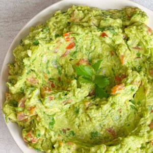 A bowl of creamy mashed avocadoes mixed with slices of tomatoes and onions and parsley.
