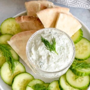 A small bowl of tzatziki sauce in a plate surrounded with cucumbers and pita bread.