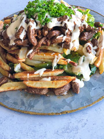 A plate of beef shawarma on top of a layer of greens an baked potatoes and topped with tahini sauce and a sprinkle of chopped parsley