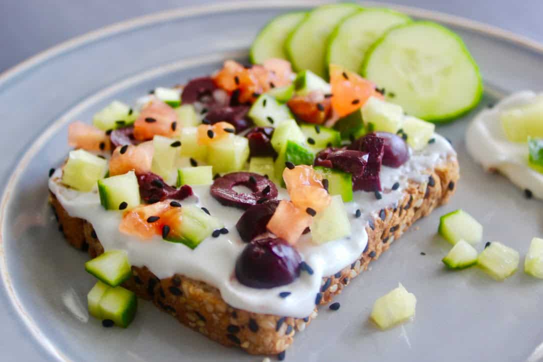 a plate with loaded toast with labneh spread topped with finely chopped Kalamata olives, cucumbers, and tomatoes