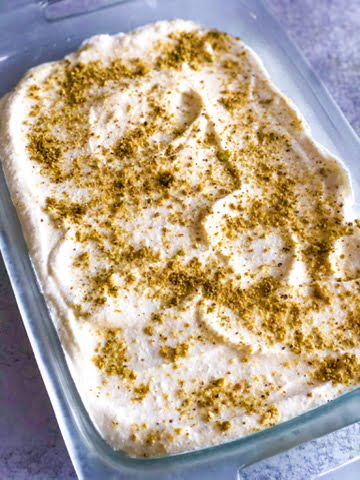 a pan of rice and cheese cream layered dessert of rice, cheese and ashta topped with ground pistachios