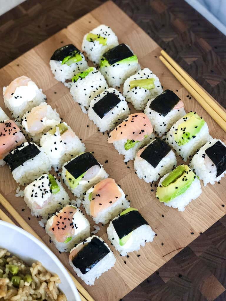 A wooden plate of No-roll sushi cubes made with short grain rice, avocado slices, asparagus, cooked shrimp, sushi nori, smoked salmon and decorated with Nigella seeds