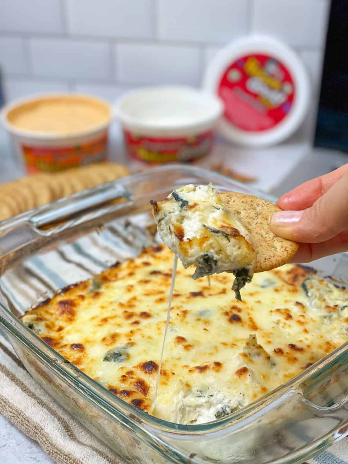 An image showing a delicious casserole of Spinach Artichoke Dip. A hand holding a cracker pulling some of the dip, creating a cheese pull.