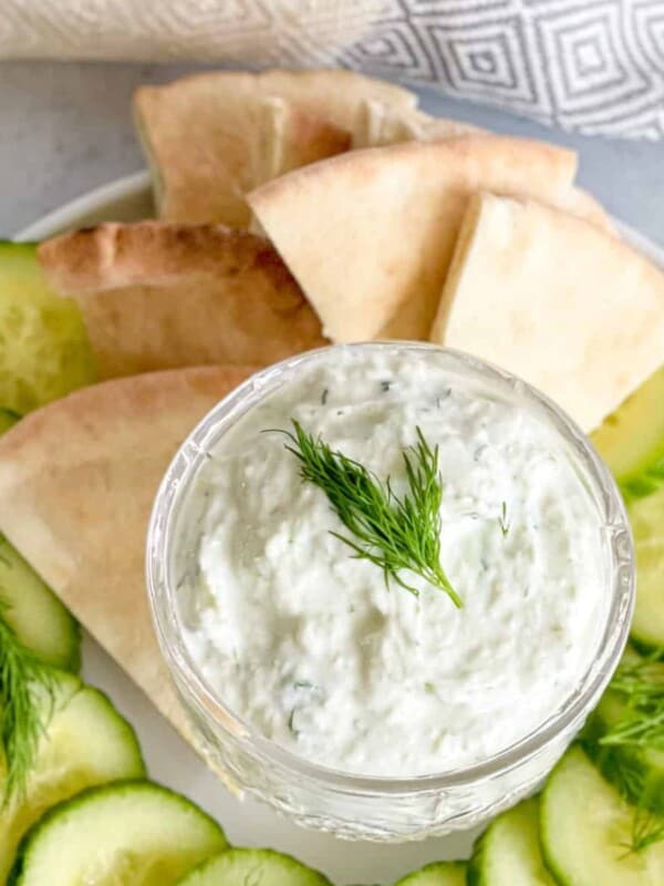 A plate featuring cucumbers and pita bread triangles with a small bowl in the middle filled with homemade tzatziki dip.