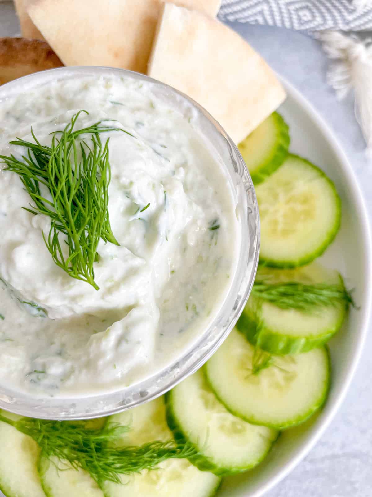 A small bowl of tzatziki sauce in a plate surrounded with cucumbers and pita bread.