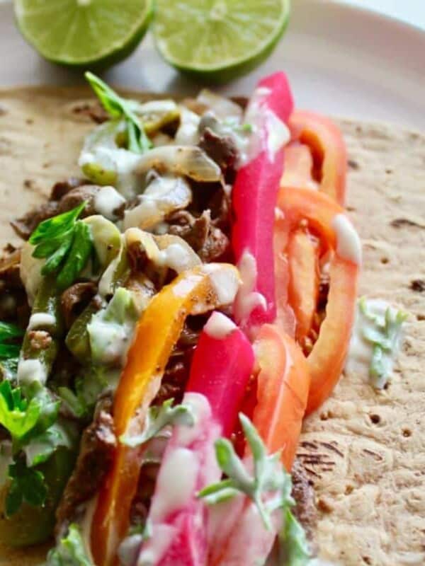 beef shawarma, pickles, chopped parsley, tomatoes, and a drizzle of tahini sauce are placed on top of a pita bread.