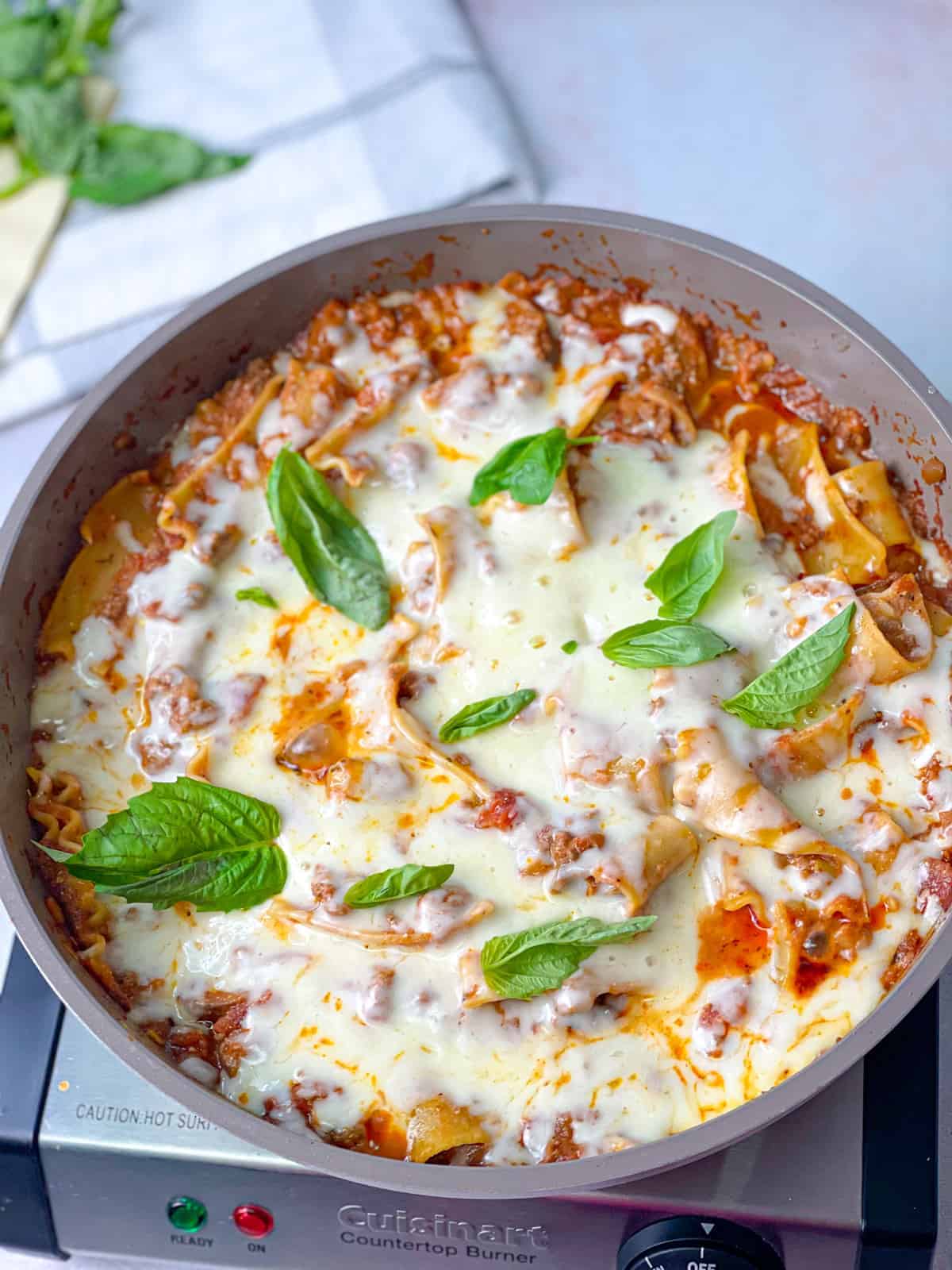 A cooking pan on a stove filled with lasagna pasta with red sauce topped with cheese and fresh green basil.