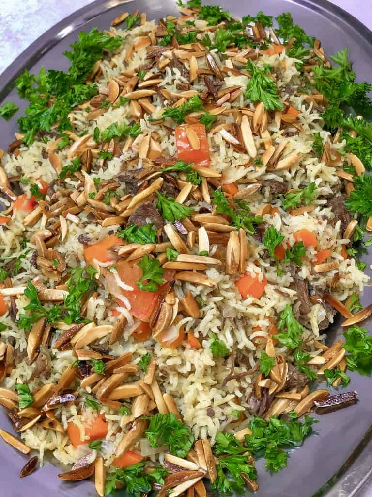 In a black plate, some brown nuts, diced vegetables, and chopped parsley top beef shanks. Under the meat pieces, cooked basmati rice sits forming with all others kabsa wirt beef dish.