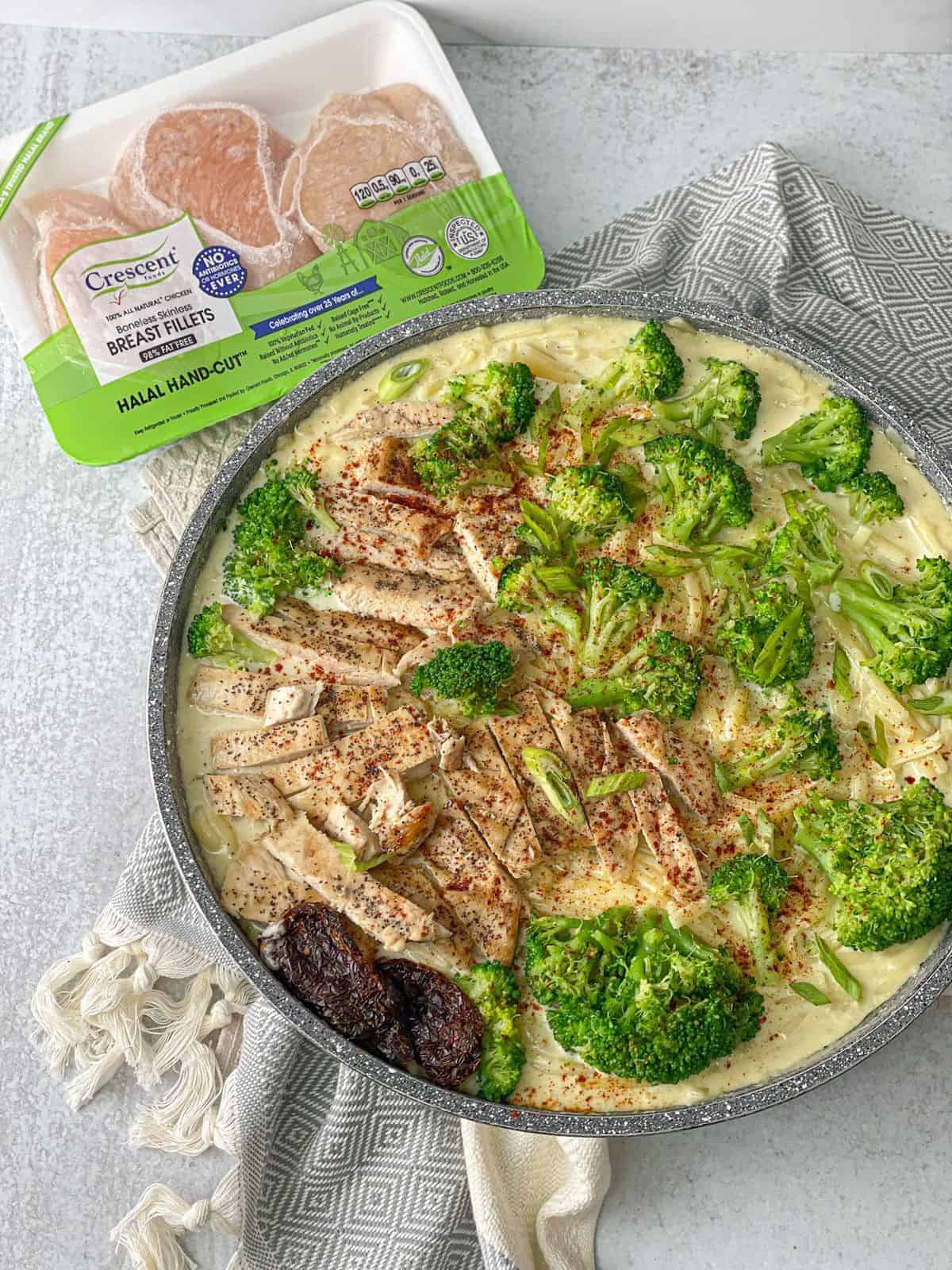 a pan of Chicken Fettuccine Alfredo with broccoli florets and grated parmesan next to a plate of breast fillets