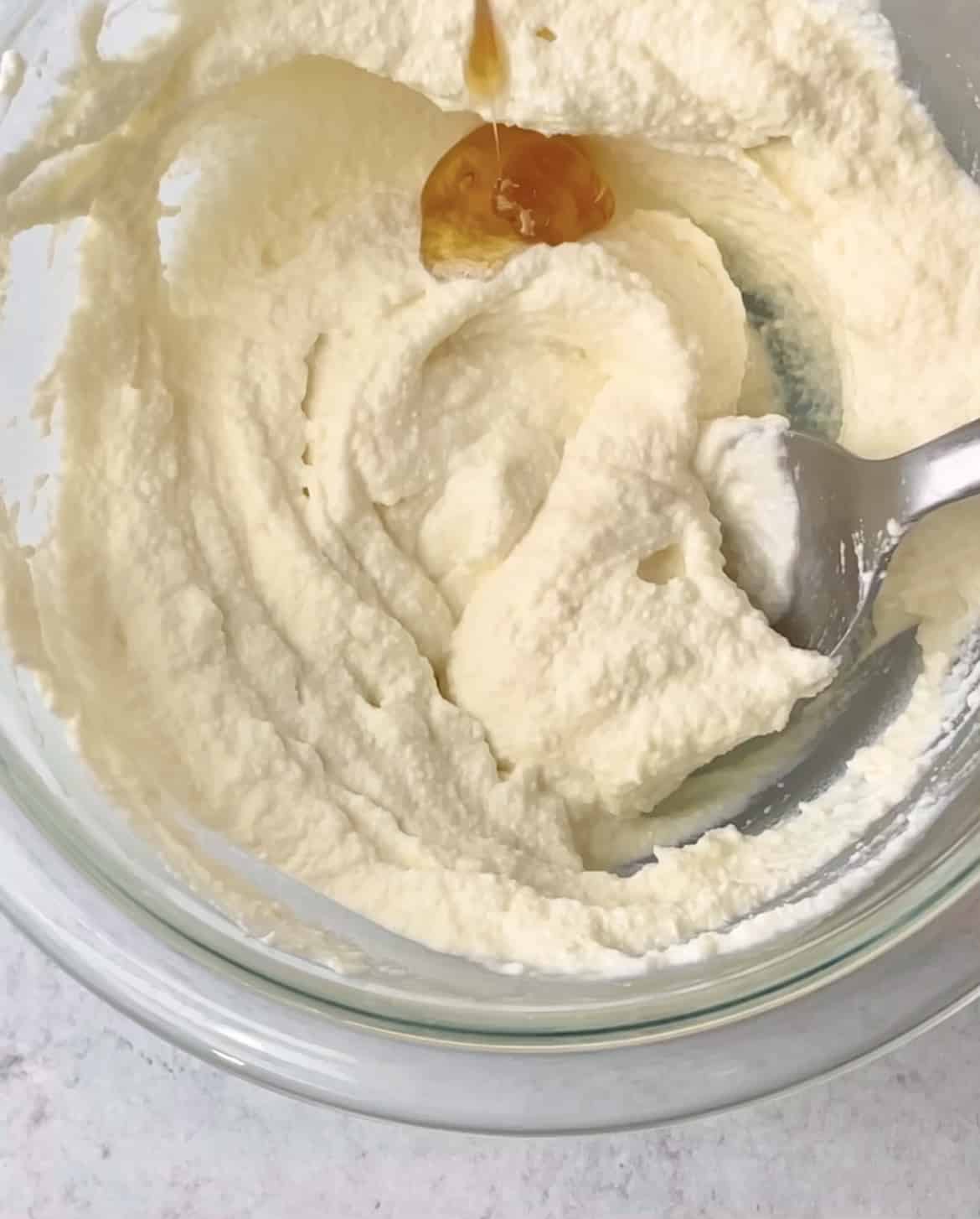 Whipped ricotta cheese is mixed with a spoon of honey in a bowl.
