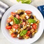 A plate of Mediterranean salad made with rotini, feta cheese, olives, and tomatoes topped with oregano and basil leaves