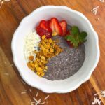 Top shot of chia seed pudding topped with sliced strawberries, granola, and shredded coconut in a white bowl on a wooden board.