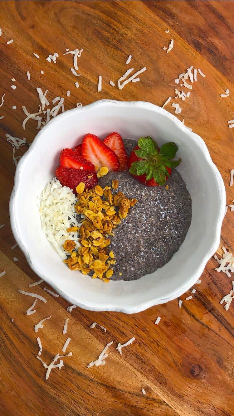Chia Seed Pudding Chia seed pudding topped with sliced strawberries, granola, and shredded coconut in a white bowl on a wooden board.