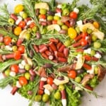 A festive Christmas wreath board arrangement of cured meats, cheeses, some veggies, and herbs to give it a wreath-like appearance. Some pine cones and decorative berries place nearby.