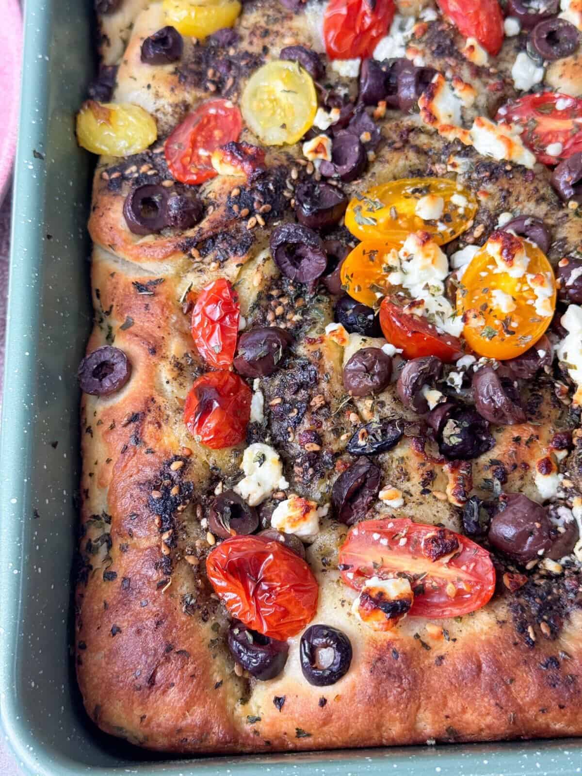 Warm, herb-infused focaccia in a pan, featuring a golden crust, zaatar drizzle, and a colorful mix of olives, tomatoes, and feta.
