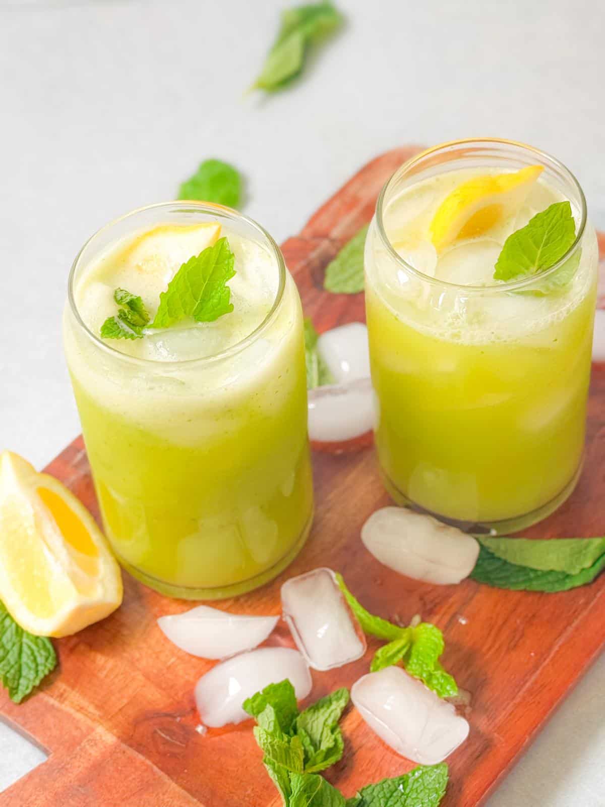 two cups of mint lemonade with lemon and mint garnish served on a wooden platter with ice, mint, an lemon wedges