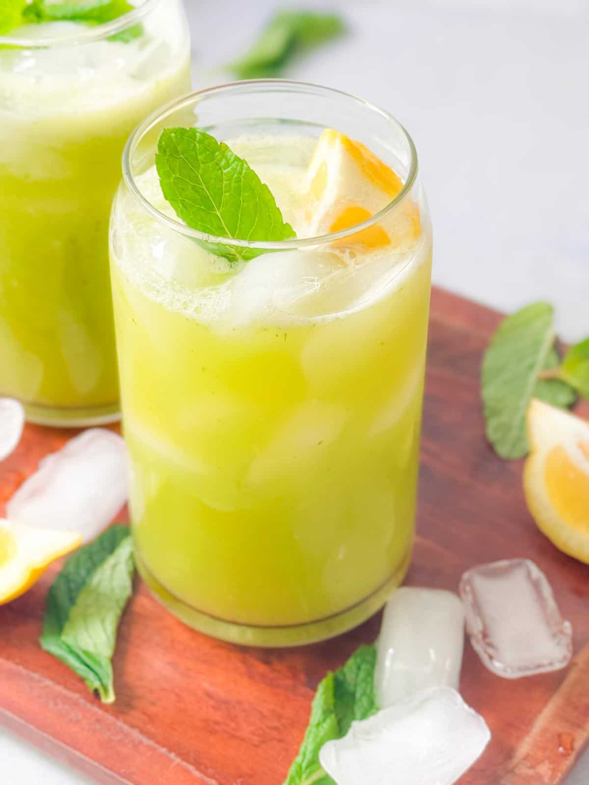Authentic Mint Lemonade Recipe two cups of mint lemonade with lemon and mint garnish served on a wooden platter with ice, mint, an lemon wedges