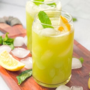 two cups of mint lemonade with lemon and mint garnish served on a wooden platter with ice, mint, an lemon wedges