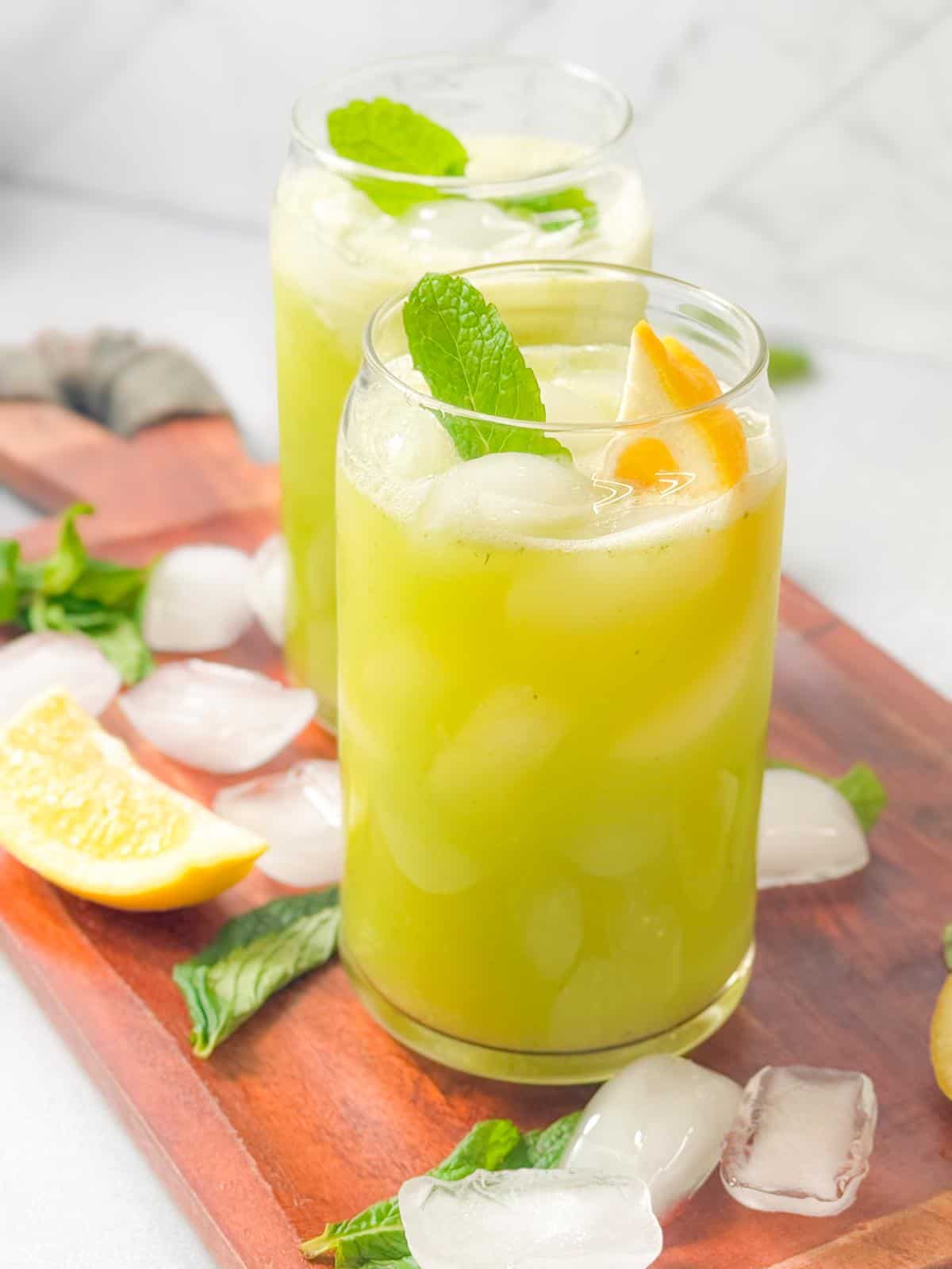 two cups of mint lemonade with lemon and mint garnish served on a wooden platter with ice, mint, an lemon wedges