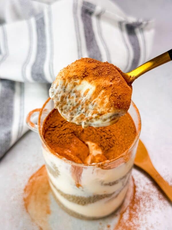 close-up of a spoon scooping creamy tiramisu chia seed pudding from a glass cup, revealing its rich layers of soaked chia seeds, coffee-infused flavor, and a dusting of cocoa powder on top