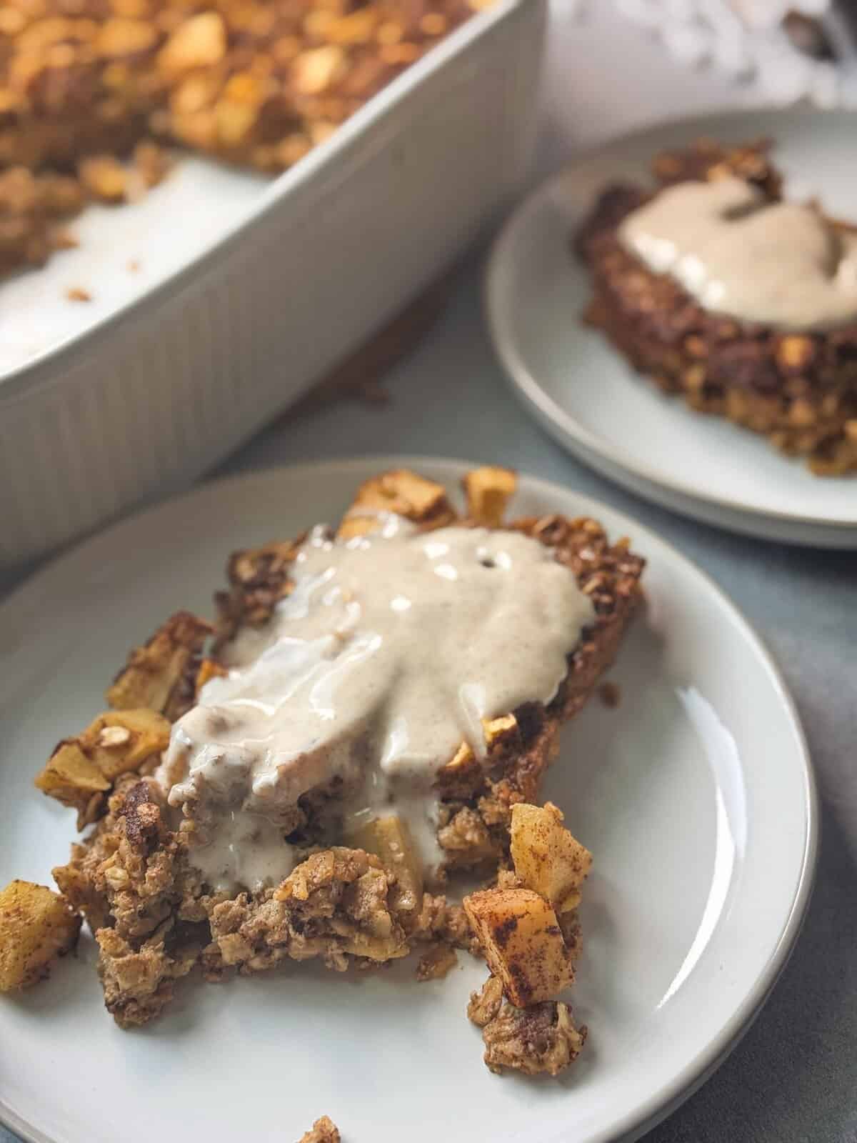 Apple Cinnamon Baked Oatmeal two slices of cinnamon apple baked oatmeal served with yogurt drizzle on top in two separate white plates with tray in background