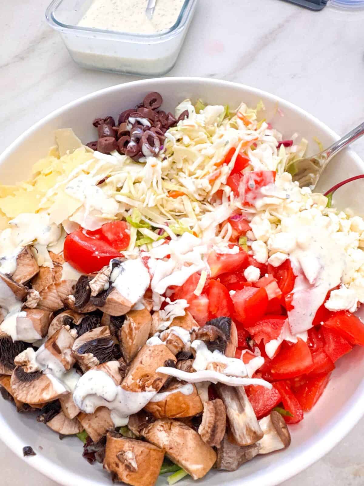 3-Ingredient Creamy Ranch Dressing Large salad bowl filled with chopped mushrooms, shredded cabbage, tomatoes, olives, and cheese, drizzled with creamy ranch dressing, with extra dressing in a glass container in the background.