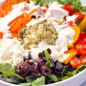 Fresh mixed salad with arugula, shredded carrots, bell peppers, feta, olives, and pumpkin seeds topped with ranch dressing in a white bowl.