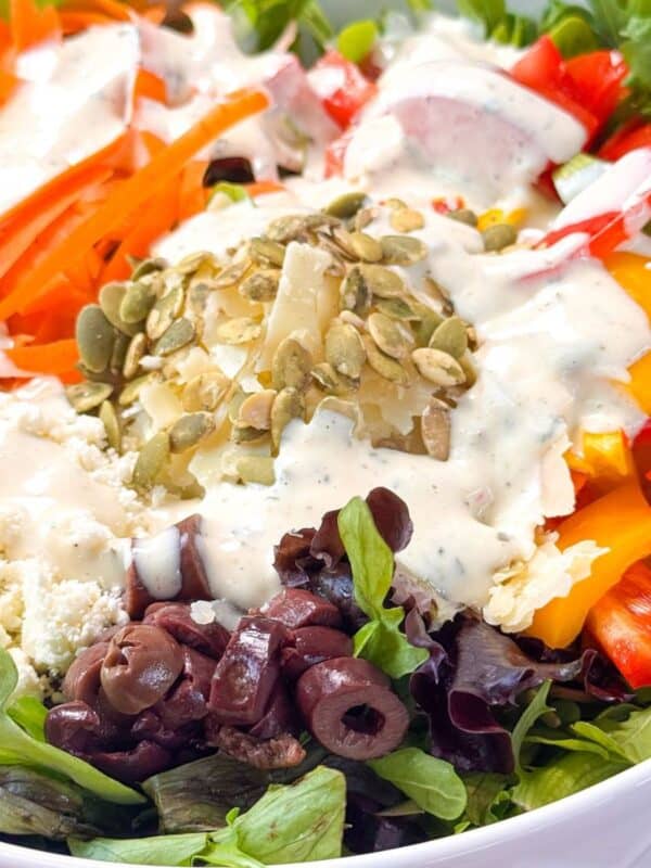 Fresh mixed salad with arugula, shredded carrots, bell peppers, feta, olives, and pumpkin seeds topped with ranch dressing in a white bowl.